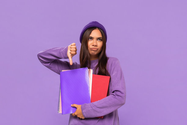 悲伤的非洲裔美国少年戴着帽子，穿着毛衣，拿着文件夹，拇指朝下，孤立地站在紫色的背景上。评价的概念。图片下载