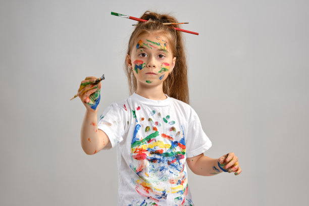 一个穿着白色t恤，头发上插着刷子的小女孩孤立地站在白布上，用涂了颜料的手和脸做手势。特写镜头图片下载