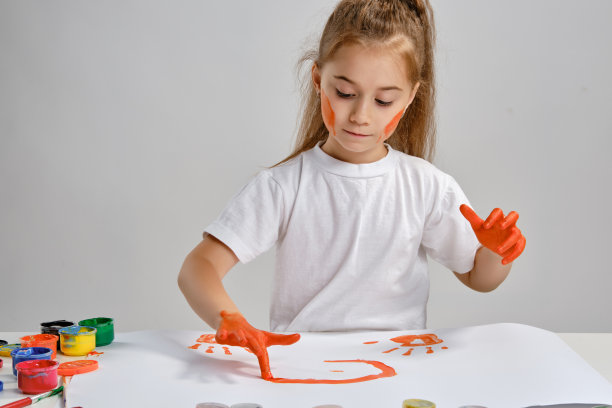 一个穿着白色t恤的小女孩坐在桌子旁边，桌上有什么人和彩色的颜料，她用手在上面画画。孤立在白色的。媒介特写图片下载