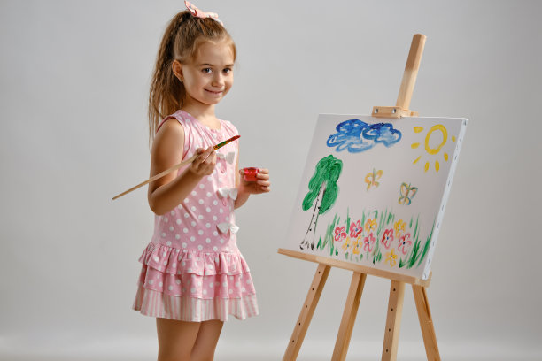 小女孩艺术家穿着粉红色的裙子站在画架后面，用画笔在画布上作画，孤立在白色的工作室背景上。媒介特写图片下载