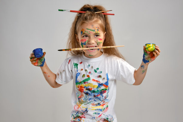 一个穿着白色t恤，头发上插着刷子的小女孩孤立地站在白布上，用涂了颜料的手和脸做手势。特写镜头图片下载