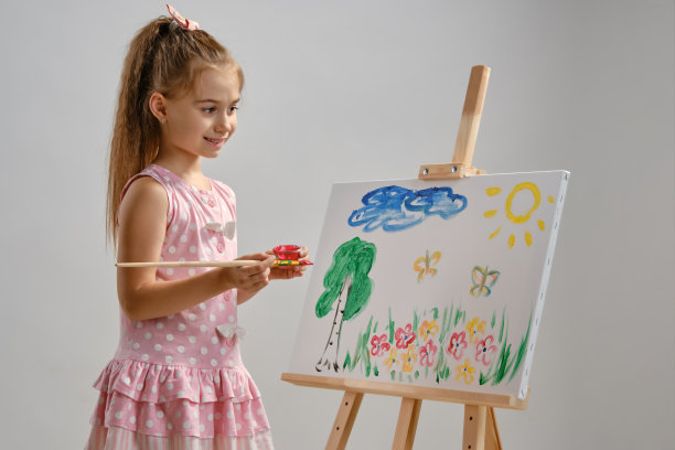 小女孩艺术家穿着粉红色的裙子站在画架后面，用画笔在画布上作画，孤立在白色的工作室背景上。媒介特写图片下载