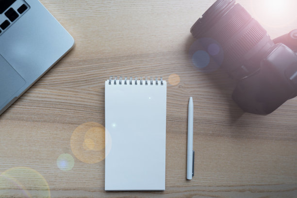 摄影师把相机放在木桌上，旁边是白纸笔记本、白笔和现代笔记本。阳光明媚的早晨，创意人员工作地点的特写图片下载