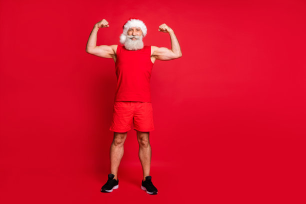全尺寸照片的快乐圣诞老人在帽子显示他的二头肌帽微笑穿着短裤运动服孤立在红色的背景图片下载