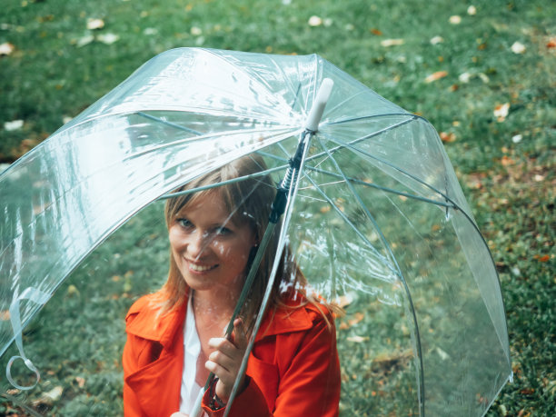 一个穿着红色华达呢的白人女孩在一把透明的伞下穿过公园。图片下载