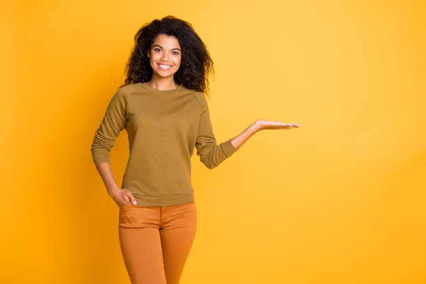 照片漂亮的黑皮肤销售员女士抱着新奇的产品在摊开的手掌提出折扣价格穿休闲套衫裤子孤立的黄色背景图片下载