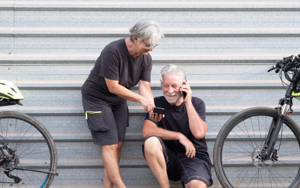 一对迷人的老年夫妇在灰色的金属面板上看着户外的手机。活动后骑电动自行车休息。健康的生活方式。黄色的头盔，黑色的衣服。图片下载