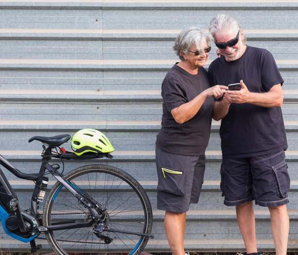 一对迷人的老年夫妇在灰色的金属面板上看着户外的手机。活动后骑电动自行车休息。健康的生活方式。黄色的头盔，黑色的衣服。图片下载