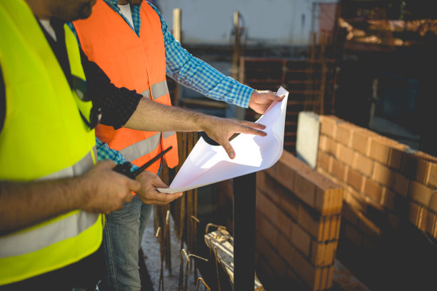 男人们戴着安全帽，穿着黄色和橙色的夹克在建筑工地摆姿势。库存图片图片下载