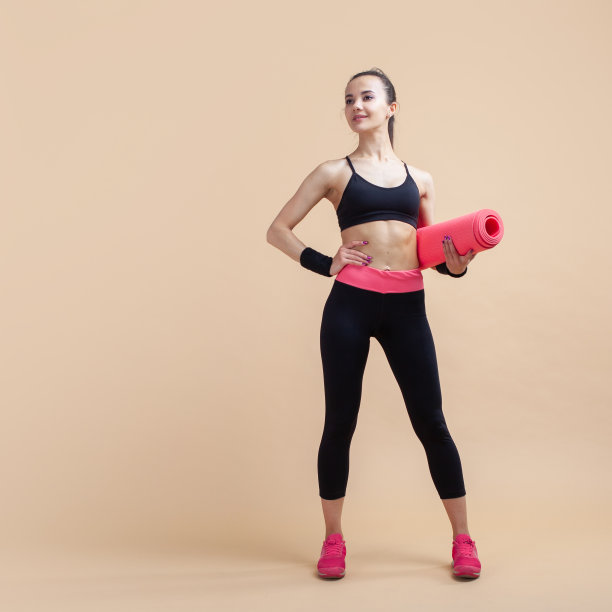 一个年轻的女孩，黑发，微笑，在粉红色的背景，在茁壮成长，板着脸，腋下挟着一个瑜伽垫。图片下载