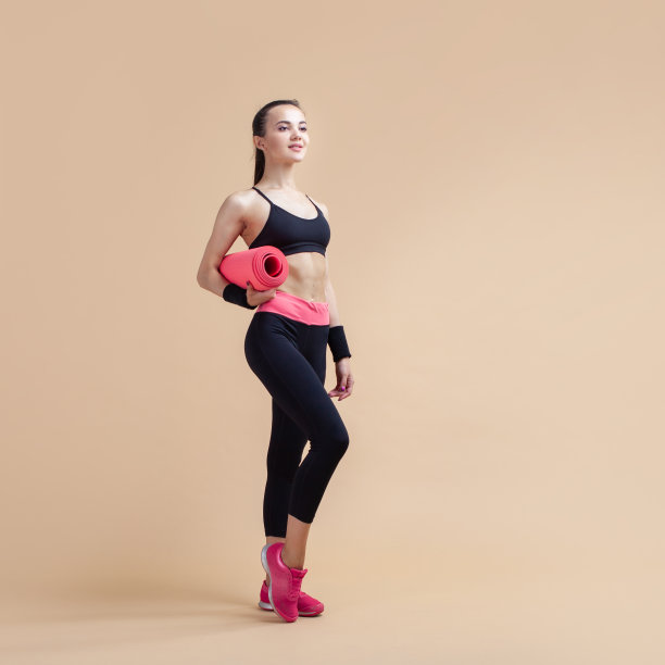 一个年轻女孩微笑着站在四分之三的地方，胳膊下夹着一个瑜伽垫。图片下载