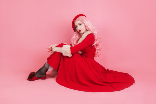 女人在红色的圣诞服装在粉红色的背景帽子。美丽的圣诞女孩拿着棒棒糖。新年的概念。漂亮的长头发模特。庆祝新的一年。甜蜜的糖果。粉色的假发。圣诞节看图片下载