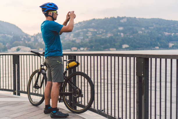 骑车者用智能手机拍照。一个戴着自行车头盔的白人男子在拍摄风景照片。户外活动。游客在意大利拍摄科莫湖图片下载