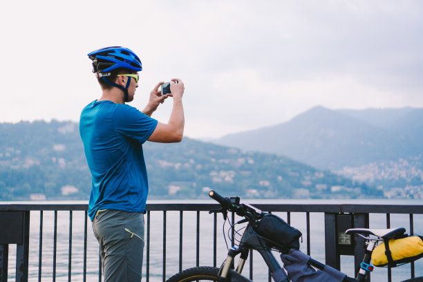 骑车者用智能手机拍照。一个戴着自行车头盔的白人男子在拍摄风景照片。户外活动。游客在意大利拍摄科莫湖图片下载