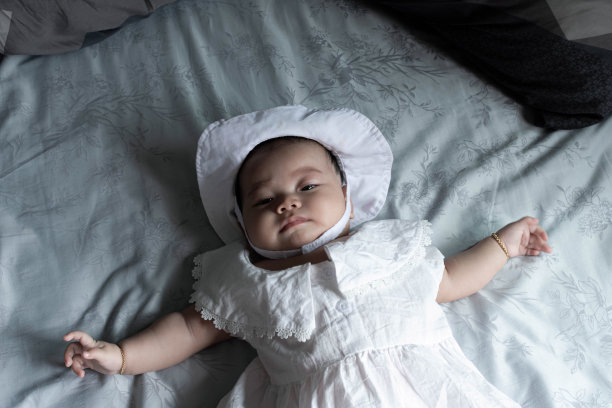 一个东南亚新生儿躺在白色的床上。东南亚的新生儿戴着白色的帽子，穿着白色的衣服。婴儿4个月大。孩子是女孩。-时尚、家庭、保健理念。图片下载