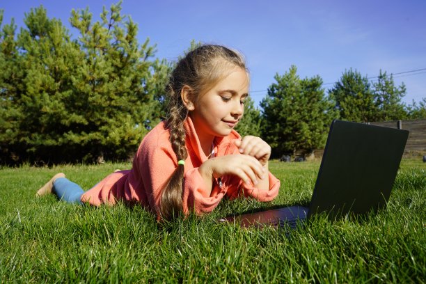 一个七岁的女孩在绿草地上用笔记本电脑工作。远程学习。一个拿着笔记本电脑的女孩，背景是松树。在街上拿着电脑的女孩。一个孩子在草地上玩电脑游戏。图片下载