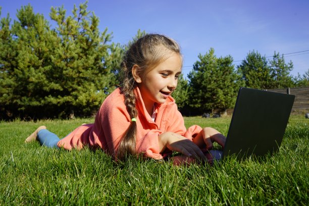 一个七岁的女孩在绿草地上用笔记本电脑工作。远程学习。一个拿着笔记本电脑的女孩，背景是松树。在街上拿着电脑的女孩。一个孩子在草地上玩电脑游戏。图片下载
