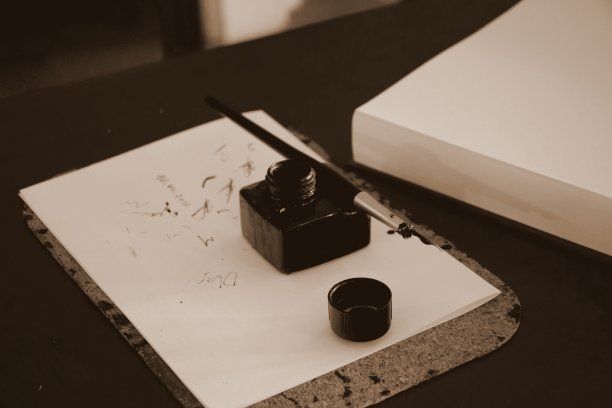 这是一张墨水瓶和羽毛笔的照片，盖子没有盖，放在一张木桌上写了一半的纸上，旁边是波兰室内的一本大书或一堆纸图片下载