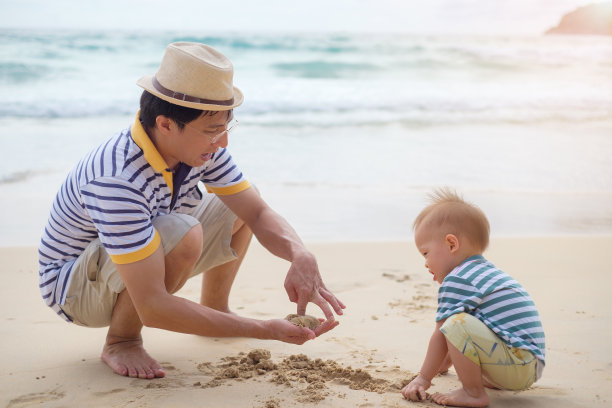 幸福的家庭。父亲和可爱微笑的18个月大的亚洲小男孩在户外白色沙滩上玩沙子，图片下载
