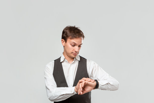 穿着白衬衫和灰色背心的年轻商人正在看他的手表。守时的概念图片下载