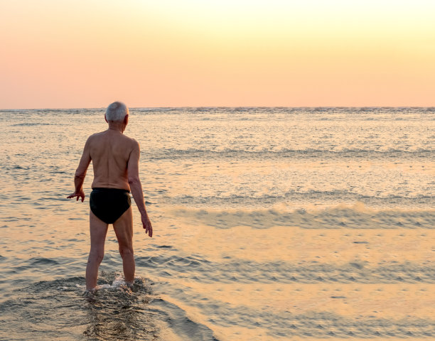 体格健康的老年男性，穿着内裤，在海滩度假享受退休后的生活，兴奋地在日出/日落的黄金时间泡在大海/海洋里。图片下载