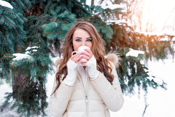 冬天里，在白雪和圣诞树的背景下，一个留着长发的女孩从马克杯里喝着热饮咖啡茶。暖和的夹克和白色毛衣。周末冬季度假的概念。图片下载