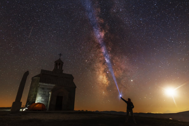 美丽的夜晚景观。山上的小教堂。教堂后面有明亮的银河和月亮。人的剪影用手电筒照亮星空。图片下载
