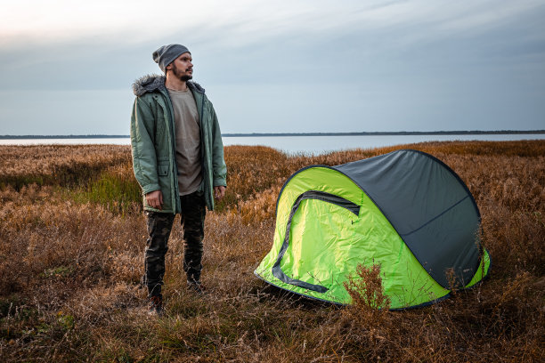 一个留着胡子的男人站在绿色的露营帐篷旁，以自然和湖泊为背景。旅游、旅游、露营的概念。图片下载