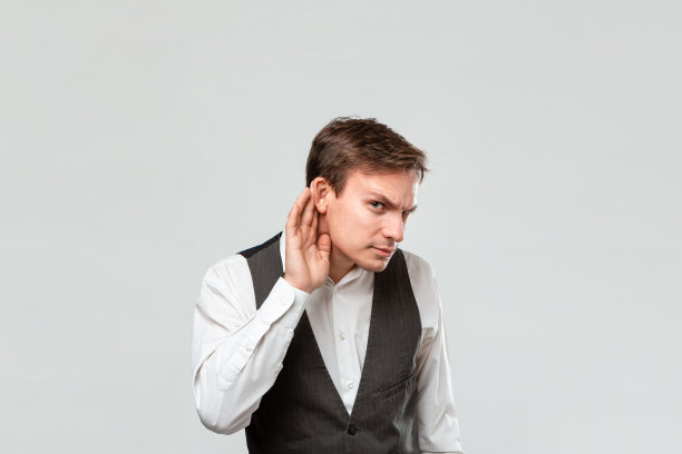 英俊的年轻人在白衬衫和灰色背心手靠近耳朵听一些有趣的谣言。图片下载