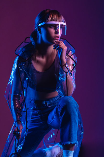 时尚迷人的年轻女子性感的20多岁的女孩模特穿着时尚时尚的透明雨衣未来派眼镜看着远处摆姿势在霓虹灯紫色工作室背景，垂直照片图片下载