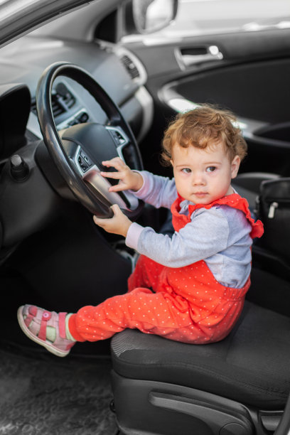 小女孩在开车。一个1岁的孩子穿着红色连身裤坐在驾驶座上。图片下载