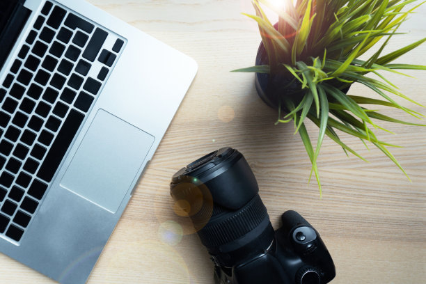 摄影师把相机放在一张木桌上，旁边放着绿色的花和现代的笔记本电脑。创意人员工作场所特写。图片下载