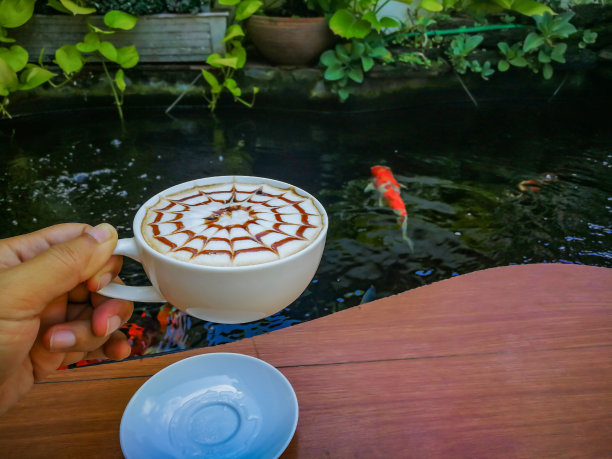 在鱼塘边，人们手捧一杯巧克力酱和焦糖酱拉花咖啡。图片下载