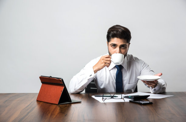 印度商人在工作/办公室喝咖啡/茶，坐在办公桌或桌子前，手里拿着茶杯和茶托图片下载