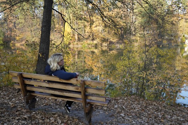 一个女孩坐在长凳上，背景是湖泊和秋天的森林。女孩在湖边的秋日森林里的长椅上休息。图片下载