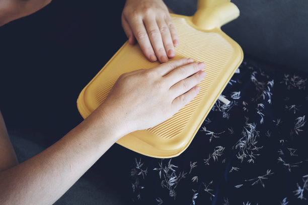 年轻女子躺在沙发上，肚子上抱着黄色的热水袋图片下载