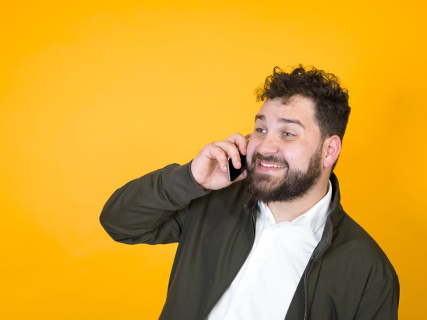 黑胡子黑头发的酷男人拿着智能手机在橙色背景前摆造型图片下载