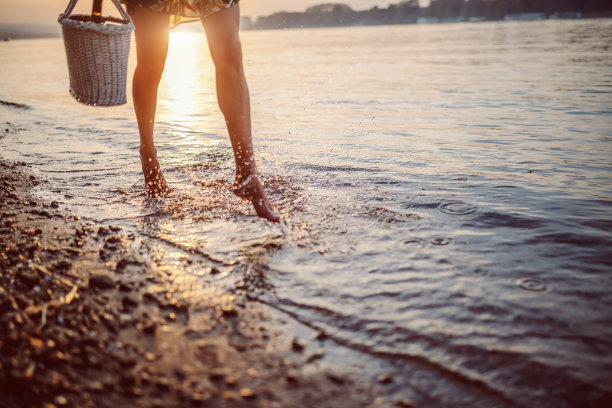 裁剪照片的女性的腿走在水里。手里拿着野餐篮子的女人。背景是太阳。图片下载