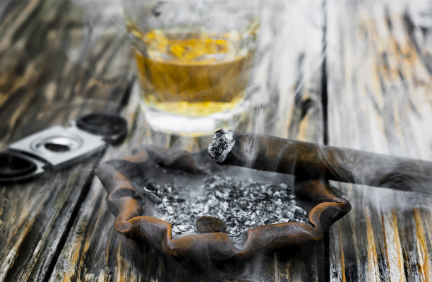 一张旧木桌，烟灰缸上放着一支还在冒烟的雪茄。背景是一根烟，一个离焦的断头台和一杯威士忌图片下载