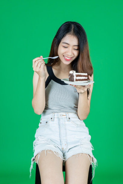 快乐的年轻亚洲女孩吃巧克力蛋糕在绿色孤立的背景。图片下载