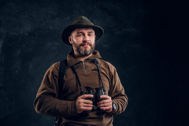 成熟的猎人拿着步枪，拿着望远镜，看着相机。暗墙背景下的工作室照片图片下载