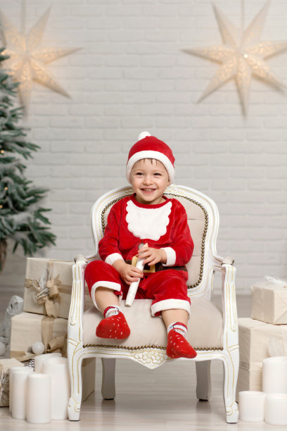 快乐的小男孩在圣诞老人服装坐在扶手椅上，拿着圣诞礼物盒图片下载