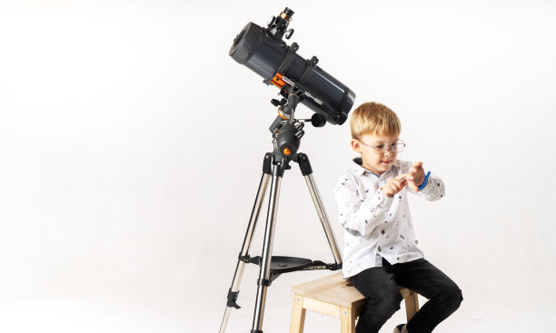 一个小男孩坐在望远镜旁边，看着他的手表，等待着天文事件的发生。图片下载