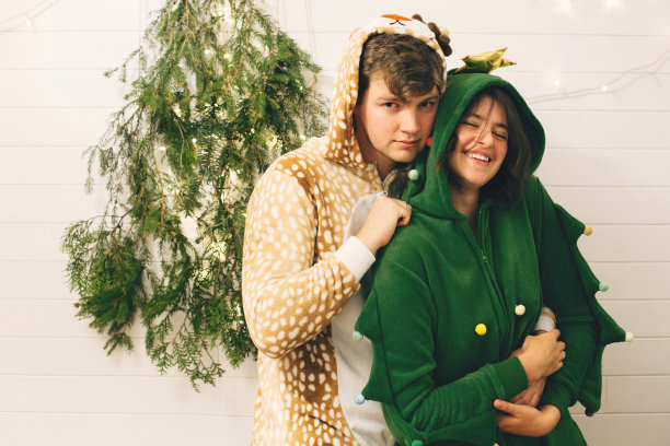 在时尚的房间里，一对幸福的情侣穿着节日睡衣在圣诞树灯前拥抱。庆祝圣诞节或新年前夕，有趣的睡衣派对。穿鹿的男人和穿圣诞树睡衣的女人图片下载