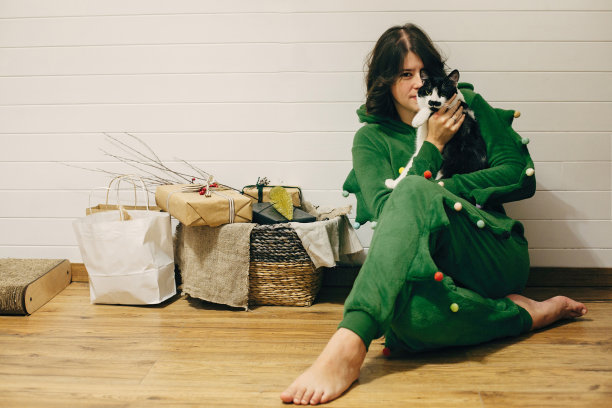快乐的女孩在节日睡衣拥抱猫，坐在地板上的圣诞礼物在时尚的房间。庆祝圣诞节或新年前夕，睡衣派对。穿着圣诞树睡衣的女人图片下载
