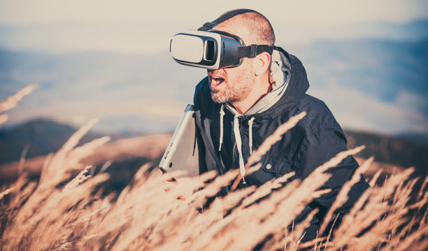 年轻人戴着VR眼镜走路。连接虚拟和现实世界的概念。胳膊下夹着笔记本电脑，戴着虚拟眼镜的千禧一代在高高的草丛中穿行。图片下载