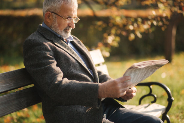 穿着灰色夹克，留着漂亮胡子的英俊爷爷坐在公园的长椅上读报纸。戴眼镜的白发老人图片下载