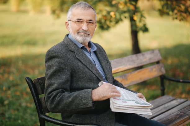 戴着眼镜的胡子老人在秋天的公园里读报纸。清晨，英俊的白发男子坐在长椅上图片下载