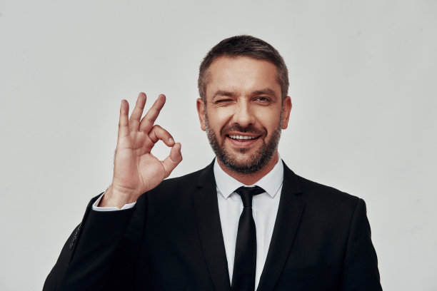 迷人的年轻男子在全套西装显示OK标志和眨眼睛，而站在灰色的背景图片下载