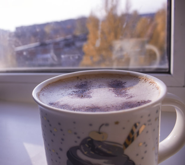 早餐喝一杯加肉桂的卡布奇诺。咖啡泡沫上的肉桂笑脸。早上在窗边喝一杯热卡布奇诺咖啡。寒冷的秋天和橙色的树叶。图片下载